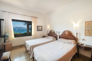 Een hotelkamer met twee bedden, een raam met uitzicht op zee, een ventilator en een nachtkastje met een lamp.