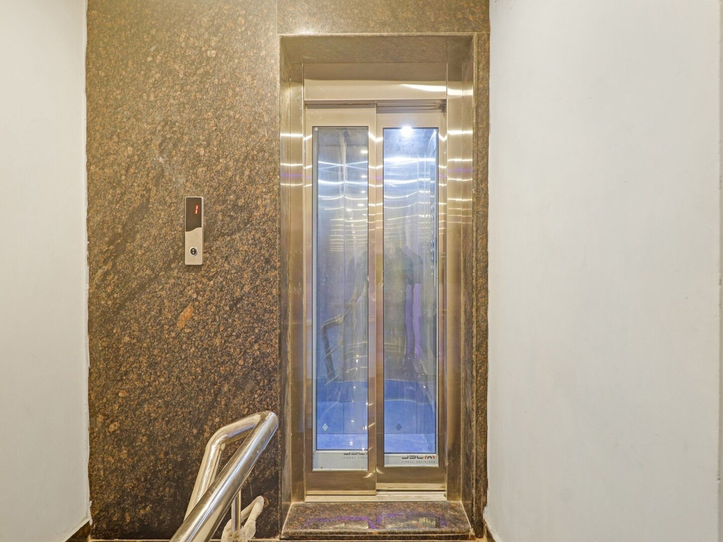 An elevator with a metallic interior and a glass door, situated in a hallway with a speckled wall.