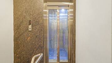 An elevator with a metallic interior and a glass door, situated in a hallway with a speckled wall.