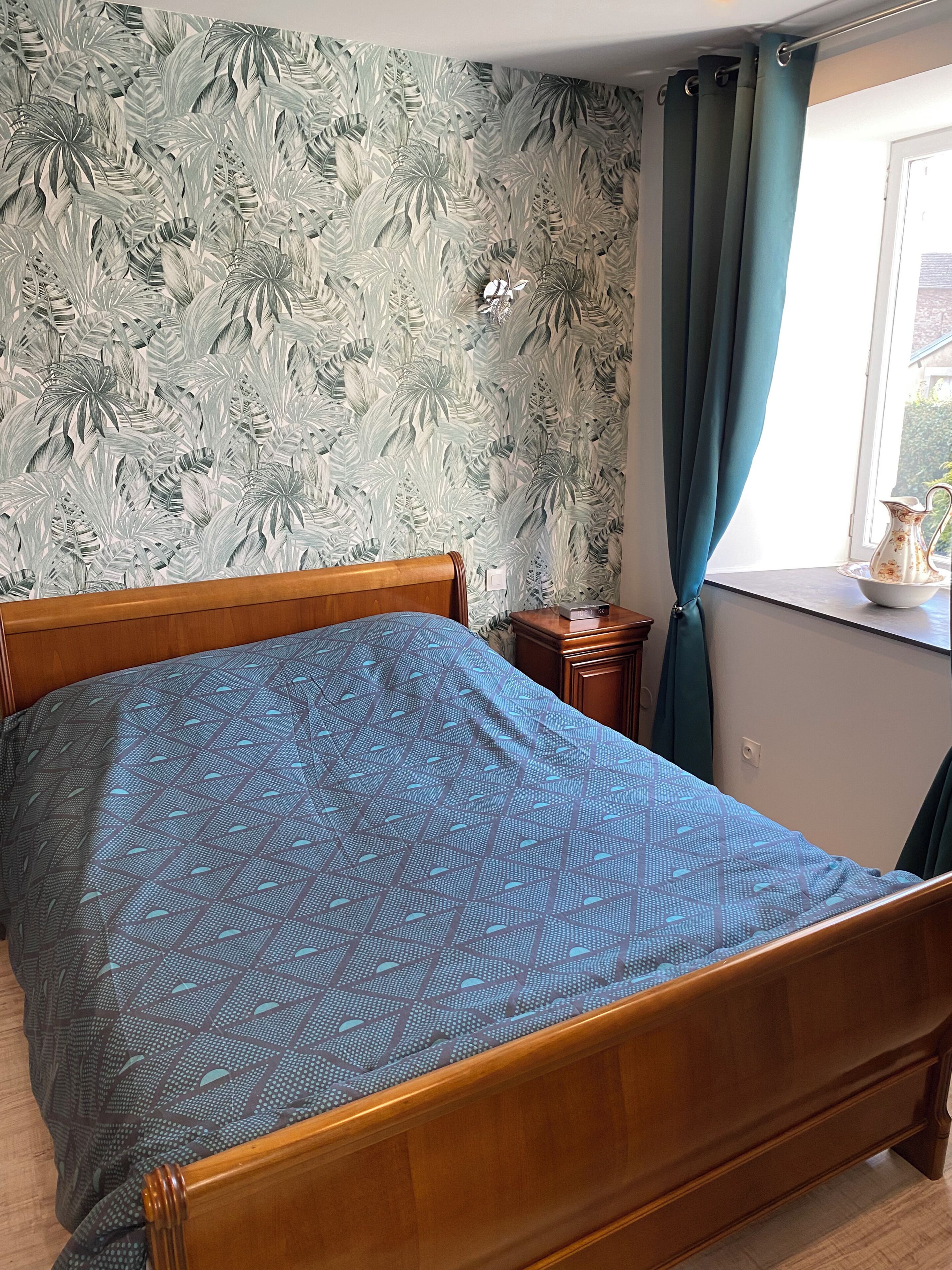 A bedroom with a bed, a nightstand, a window with curtains, and a wall with a leaf pattern.