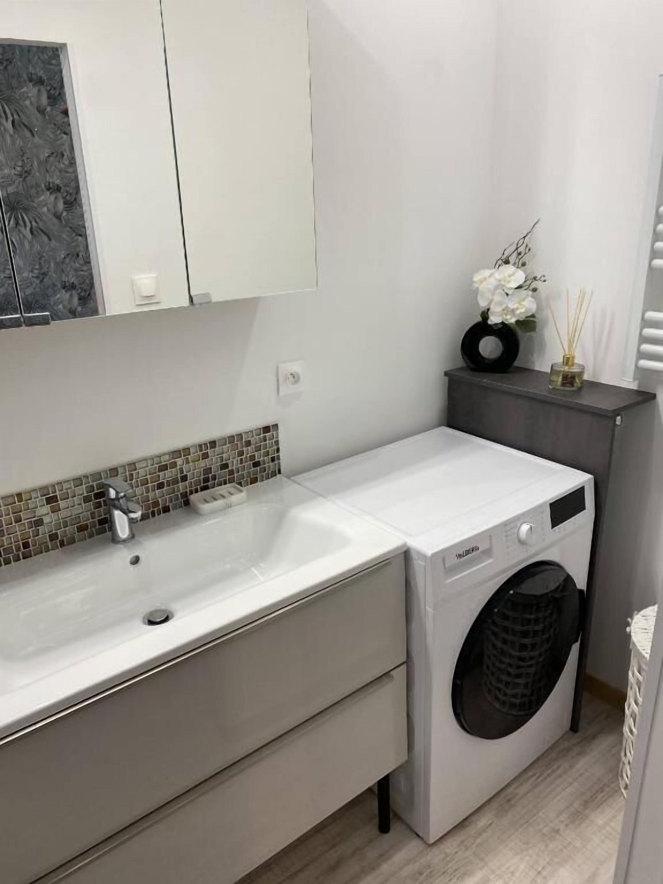 A compact bathroom with a white sink, a washing machine, and a small cabinet with a decorative item.