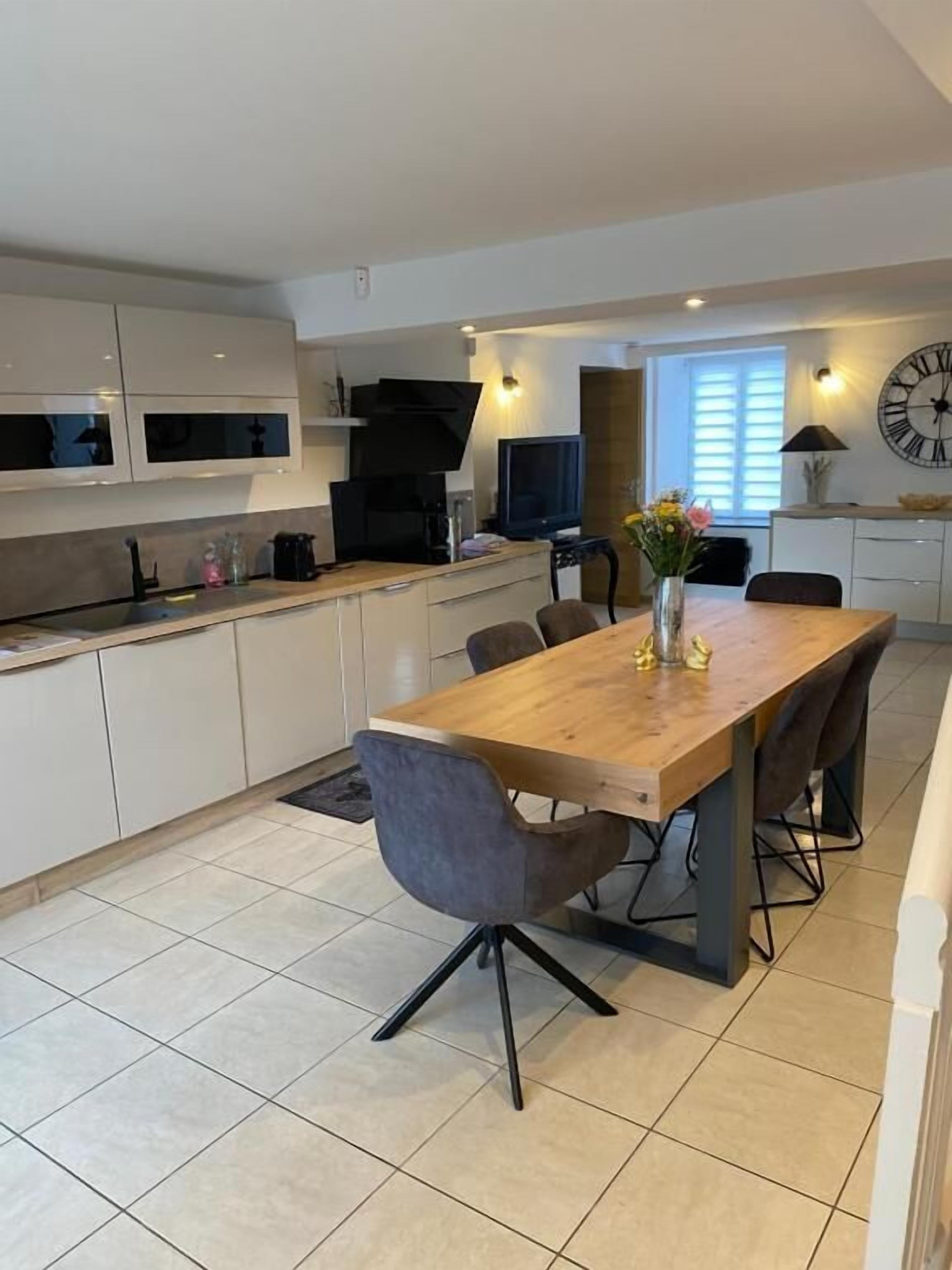 A modern kitchen with a dining area, featuring a wooden table, grey chairs, and a clock on the wall.