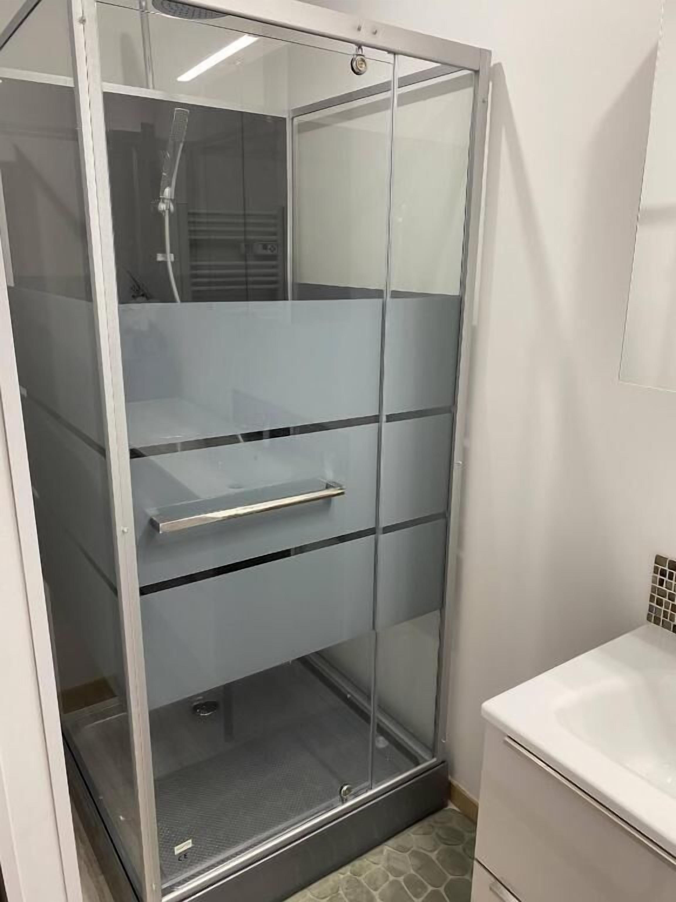 Una ducha con mampara de vidrio, un estante metálico y un lavamanos blanco con salpicadero de azulejos.