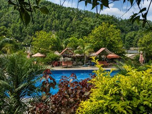 A pool area with lounge chairs and umbrellas, surrounded by lush greenery and hills in the background.
