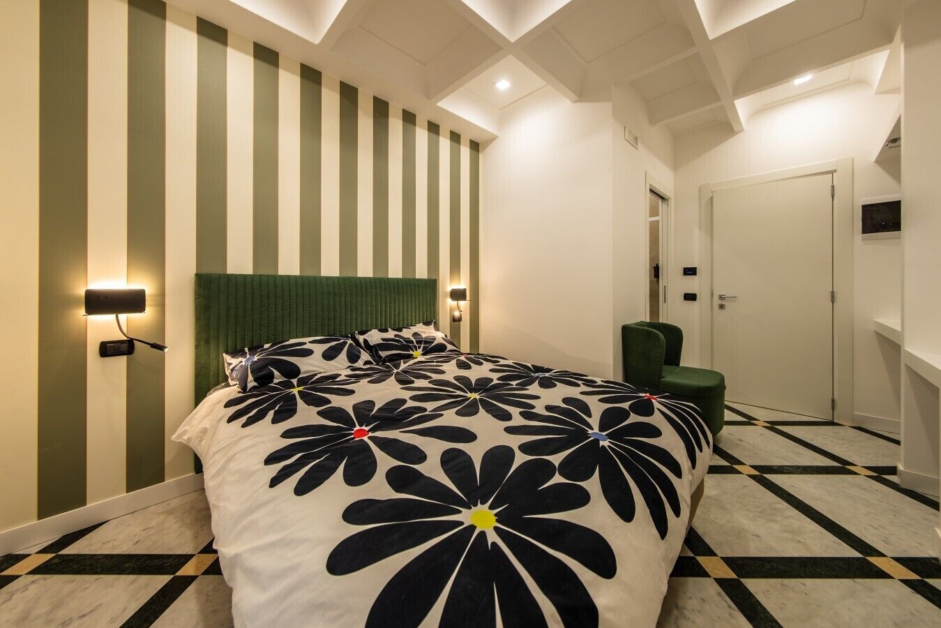Une chambre d’hôtel avec un lit, un fauteuil recouvert d’un tissu vert, une porte blanche et un sol à carreaux.