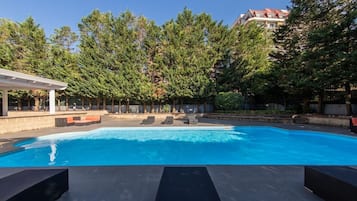 Ein Schwimmbecken mit Liegestühlen und einer überdachten Sitzgelegenheit, umgeben von Bäumen und einem Gebäude im Hintergrund.