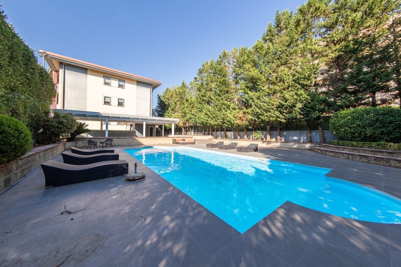 Ein Schwimmbad mit modernem Design, umgeben von einem angelegten Garten und einem Gebäude mit überdachter Terrasse.