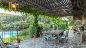 An outdoor patio with a stone floor, a wooden table, and chairs, surrounded by potted plants and a metal fence.