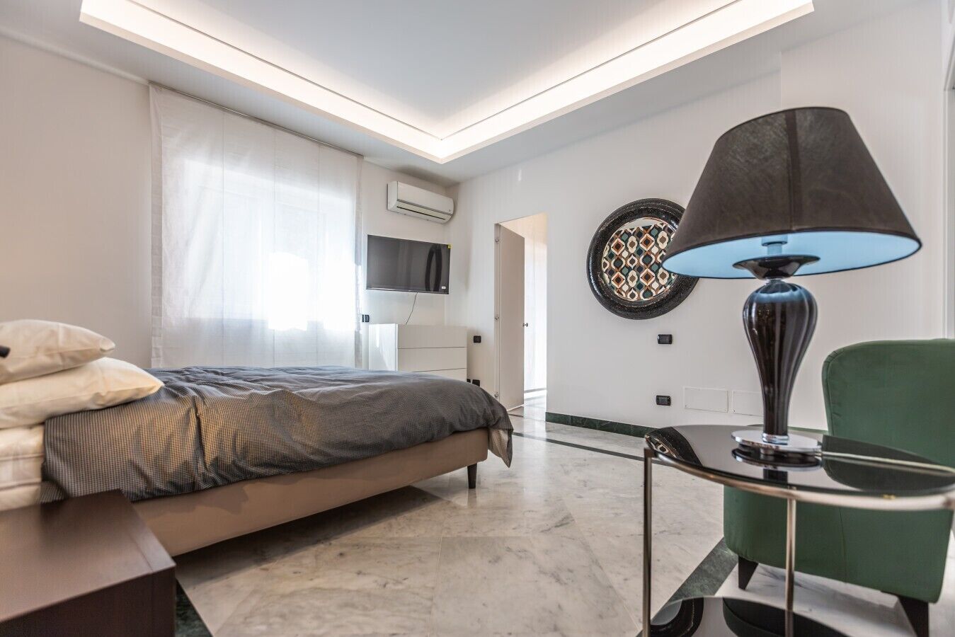 Quarto de hotel moderno com cama, escrivaninha, cadeira, luminária, televisão e um objeto decorativo na parede.