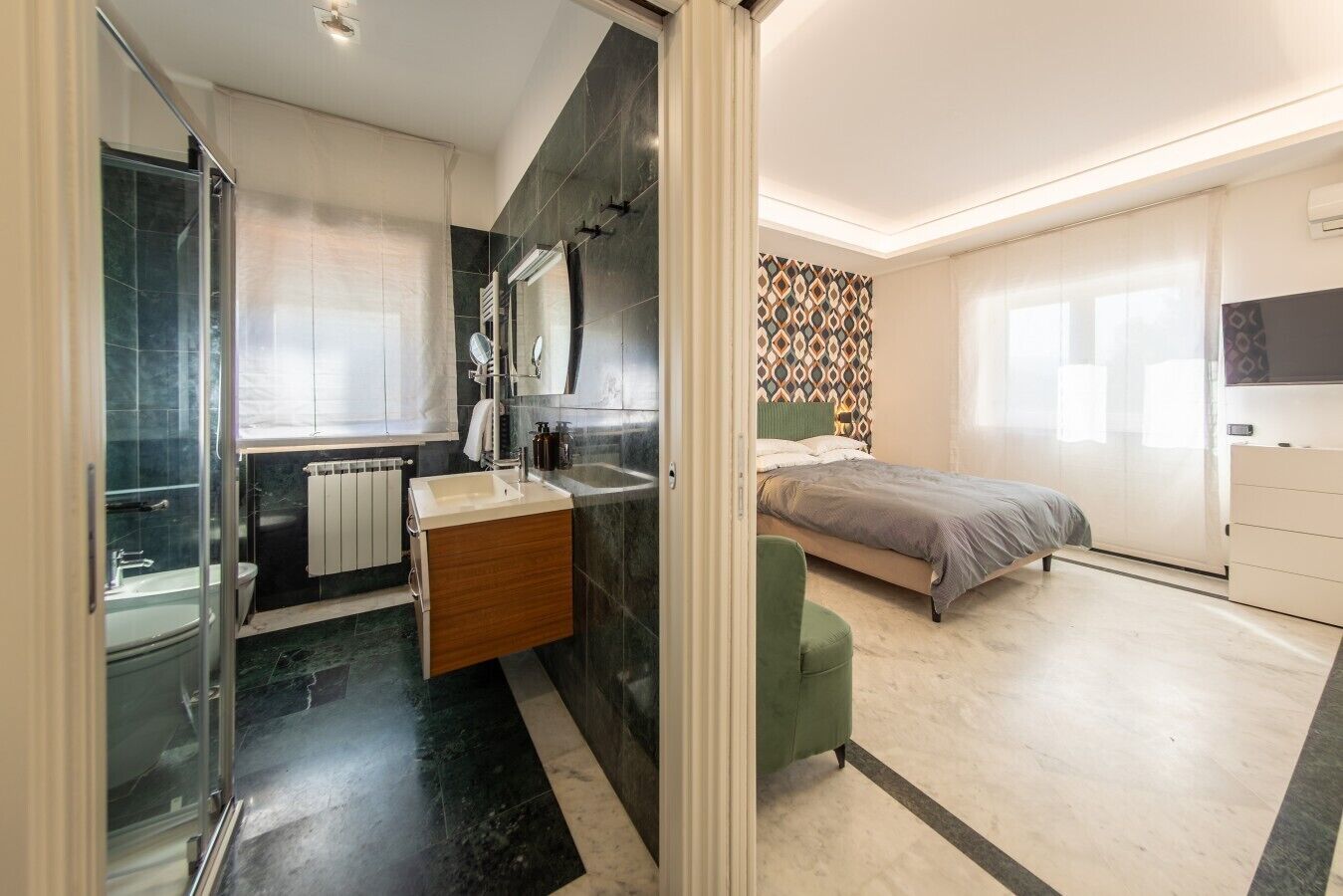 Un dormitorio moderno con cama, una silla verde, un baño con cabina de ducha de vidrio y una pared con diseño.