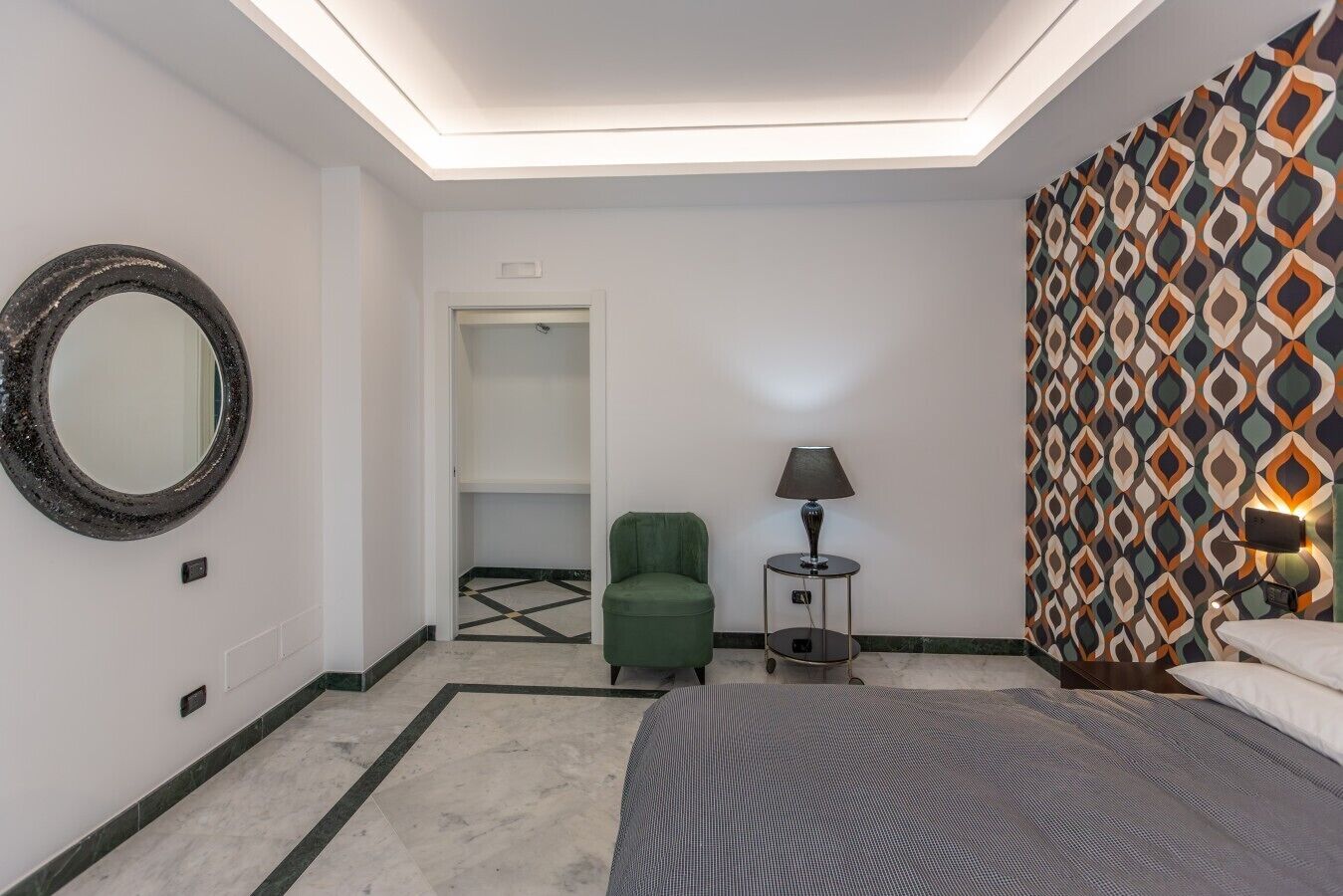 Une chambre d’hôtel avec un lit, un fauteuil vert, un miroir rond, une lampe et un mur à motifs.