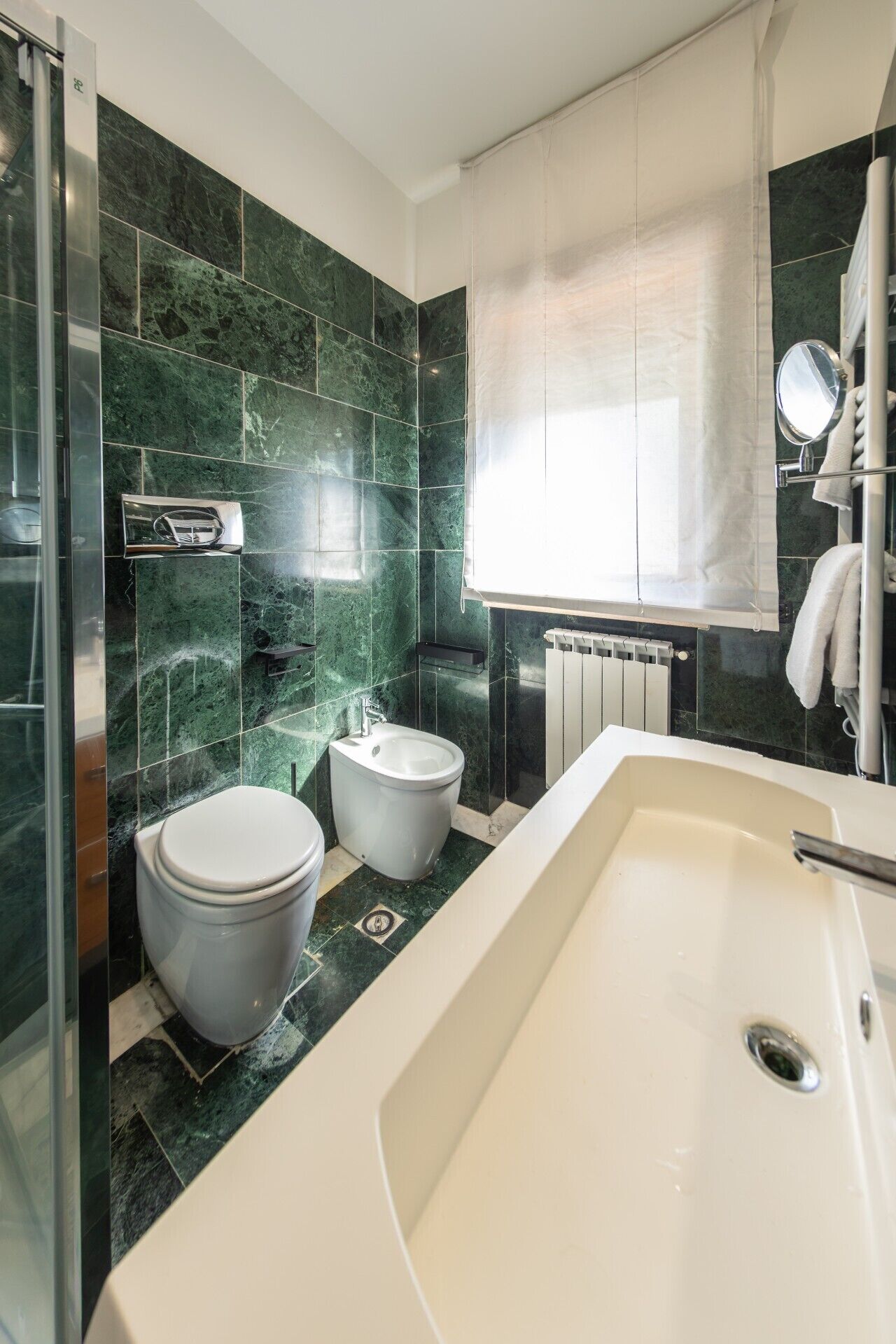 Ein modernes Badezimmer mit einer großen Badewanne, zwei Toiletten, einer Duschkabine aus Glas und einem Fenster mit weißem Vorhang.