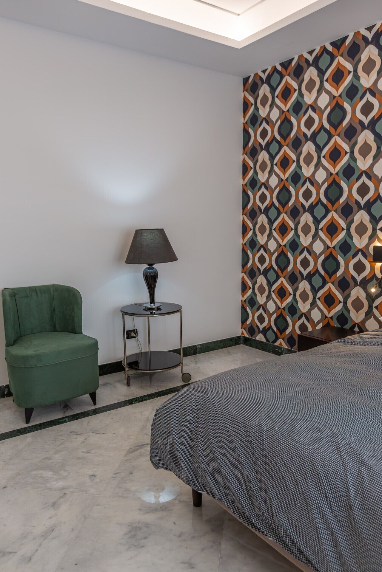A hotel room with a bed, a green armchair, a small table with a lamp, and a patterned wall.