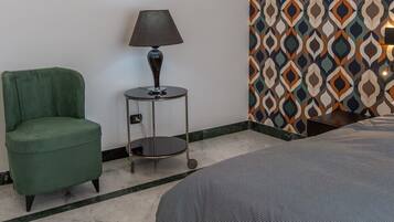 A hotel room with a bed, a green armchair, a small table with a lamp, and a patterned wall.