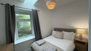 A bedroom with a bed, a bedside table, a lamp, a window with curtains, and a chandelier.