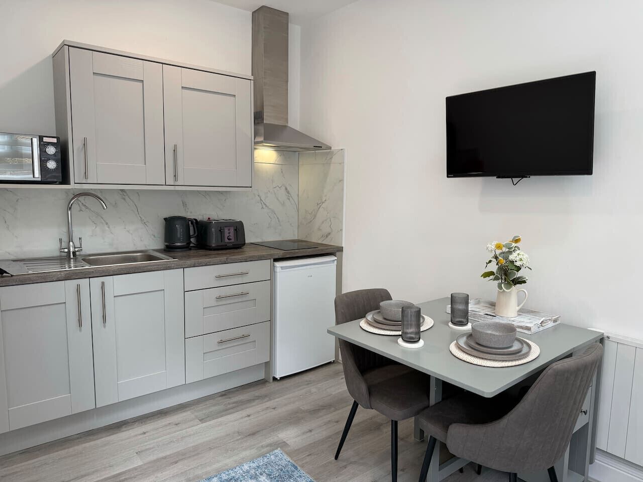A modern kitchen with a built-in microwave, toaster, and coffee maker, a dining area with a table and chairs, and a flat-screen TV mounted on the wall.