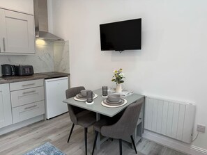 Eine moderne Küche mit integriertem Geschirrspüler, einem flachbildfernseher über der Arbeitsfläche und einem Essbereich mit zwei Stühlen und einem Tisch, gedeckt mit Tellern und einer Blumenvase.