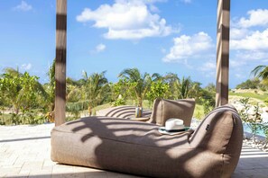 A wicker lounge chair with cushions, a remote, and a drink on a terrace with a view of palm trees and a clear sky. - Private Pool Escape - Luminara at The Reef (Sint Michiel)