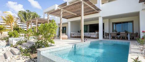 A modern outdoor pool area with a clear blue pool, wooden pergolas, and a seating area with lounge chairs.
