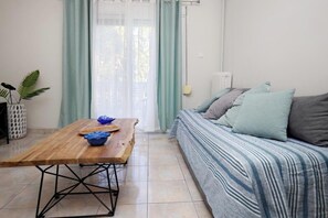 A room with a wooden table, a sofa with striped bedding, and a window with curtains. - Pantheon - A Cozy Nest in the Center of Athens (Athens)
