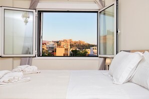 A hotel room with a bed, a window with curtains, and a view of a cityscape. - Olympus Apartment - Cozy Stay With Acropolis View (Athens)