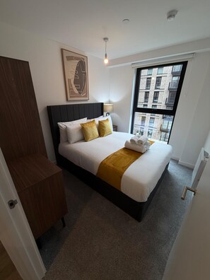 A modern hotel room with a large bed, a wooden wardrobe, a window with a city view, and a ceiling light. - Stunning 3 Bedroom Apartment-2 Bath with Balcony (Salford)