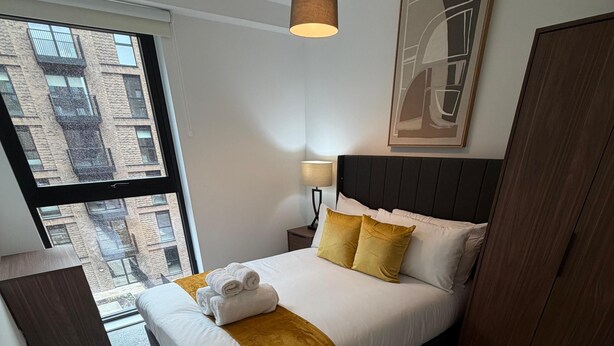 Habitación de hotel moderna con una cama grande, una mesita de noche, una lámpara y vistas a la ciudad a través de un ventanal amplio.