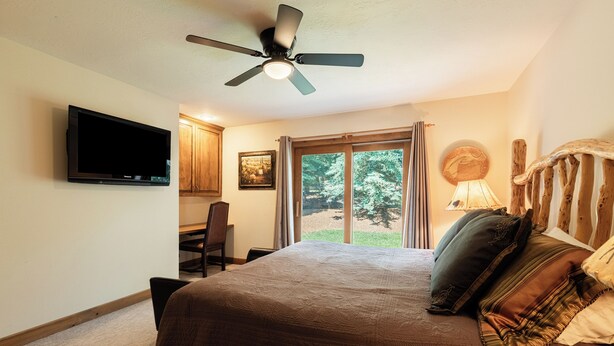 A bedroom with a bed, a ceiling fan, a TV, a desk, and a window with a view of greenery. - 6BR Deer Valley retreat with hot tub, game room, and mountain views. (Park City)