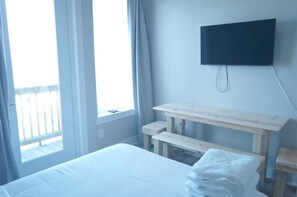 A hotel room with a bed, a wooden bench, a television mounted on the wall, and a window with curtains.