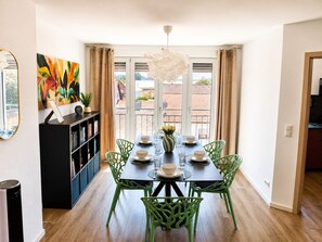 A dining area with a table set for four, surrounded by green chairs, a bookshelf, and a large window with curtains. - Premium Apartment - 90 m² - Central, near City Park (Pfaffenhofen an der Ilm)