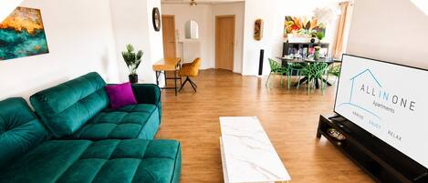 Una sala de estar moderna con un sofá verde, una mesa de centro blanca y un televisor que muestra 'ALLINONE Apartments'.