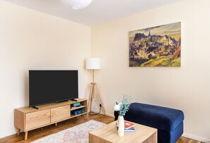 A living room with a flat-screen TV on a wooden stand, a blue sofa, a painting on the wall, and a lamp. - Edinburgh Gem Near Castle & Old Town (Edinburgh)