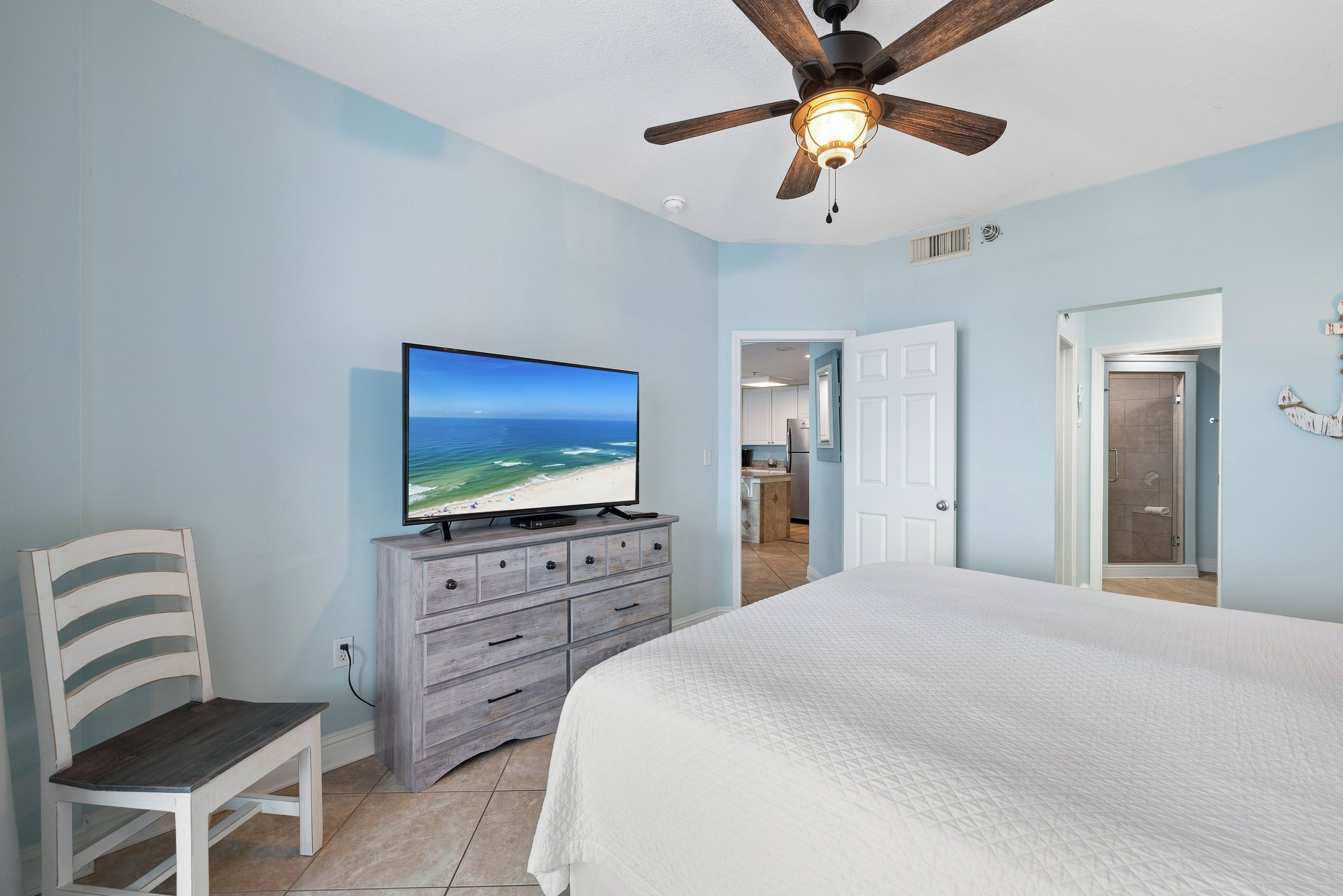 A bedroom with a large bed, a television on a dresser, a ceiling fan, and a view into the adjacent bathroom.