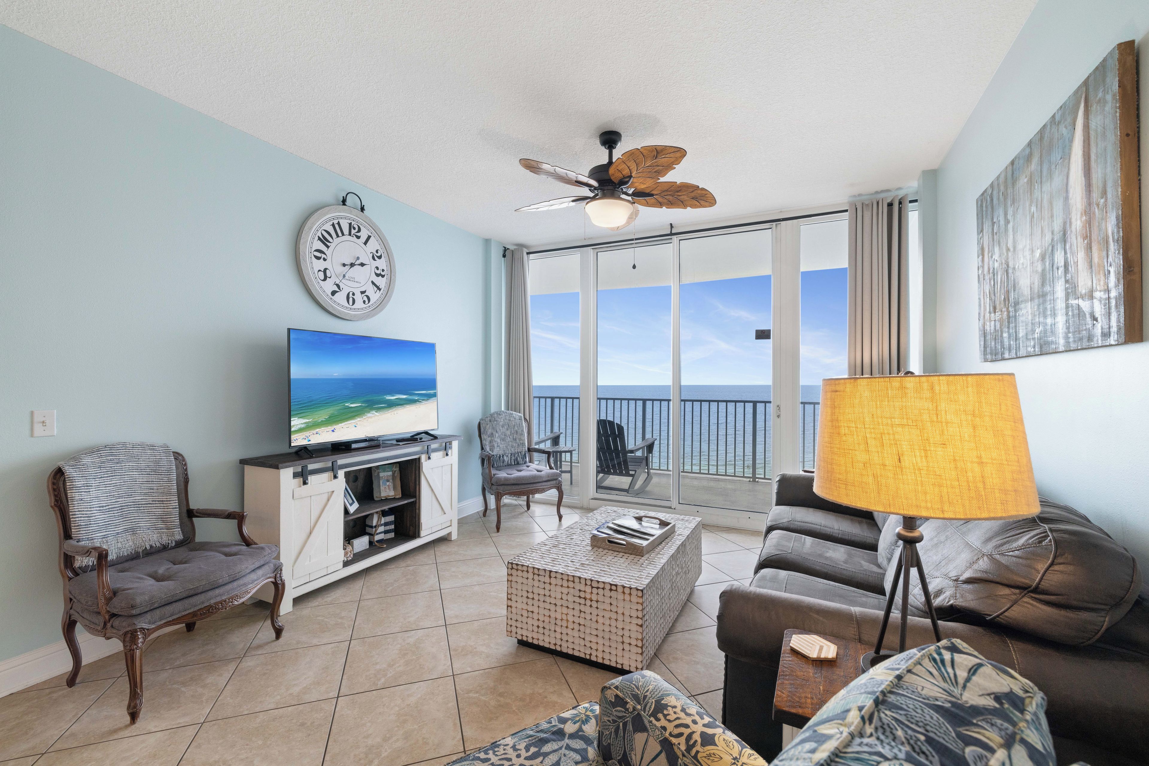 Ein Wohnzimmer mit Fernseher, einer Uhr, einem Deckenventilator und Blick auf das Meer durch große Fenster.