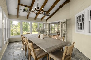 Uma sala de jantar com uma mesa grande, cadeiras, um lustre e grandes janelas.
