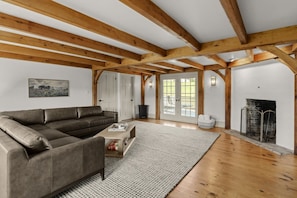 A spacious living room with a leather sofa, wooden beams, a fireplace, and a large window.