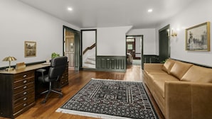 Una habitación amplia con un escritorio de madera, una silla de oficina negra y un sofá marrón. Un tapete estampado cubre el suelo, y hay una escalera que conduce a otro nivel.
