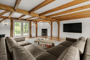 Uma sala de estar ampla com lareira, vigas de madeira, um amplo sofá e uma televisão de tela plana na parede.