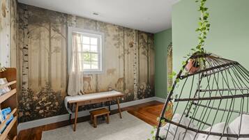 A room with a wooden floor, a window with a curtain, a bookshelf, a small wooden bench, and a hanging plant.