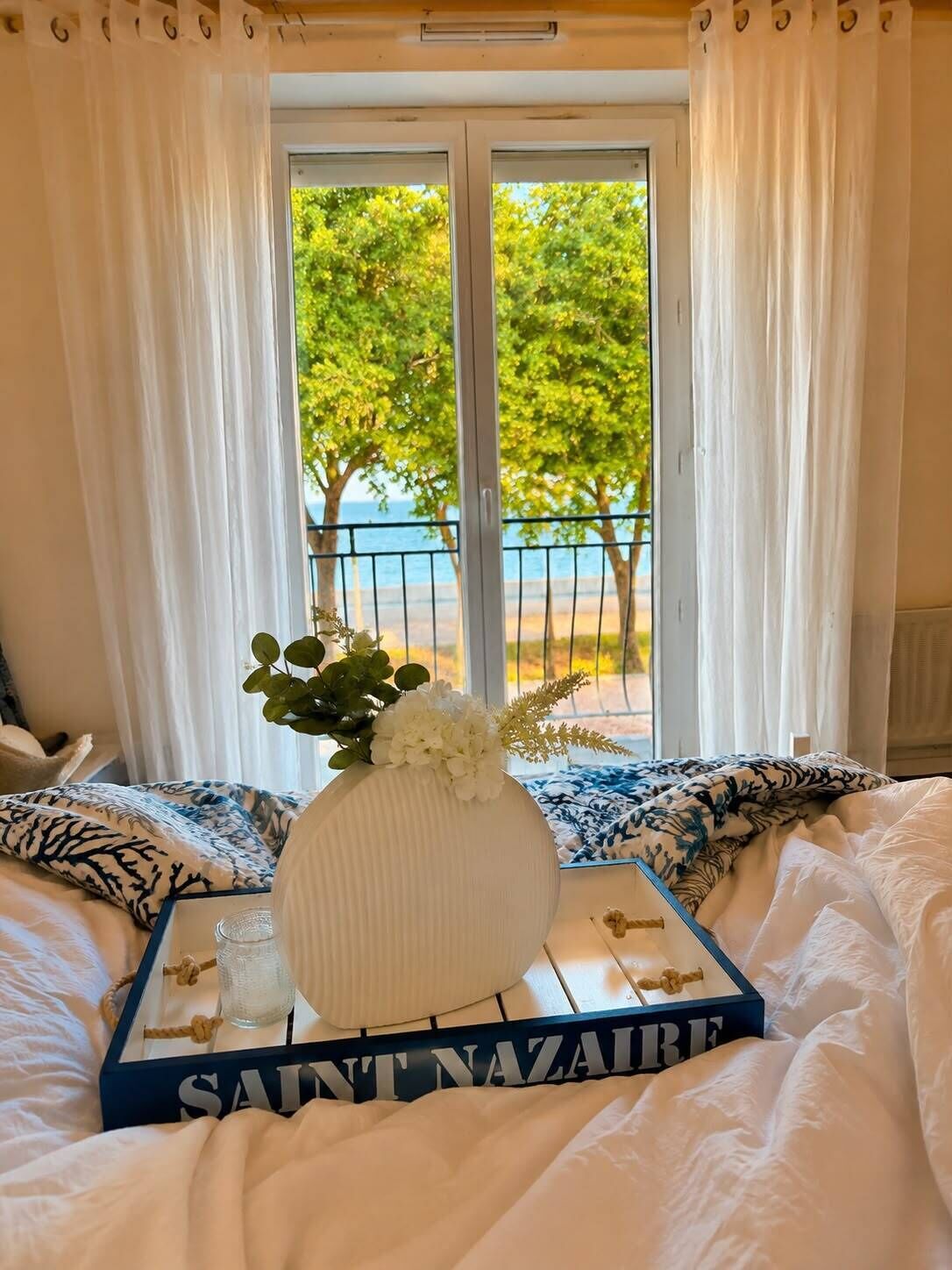 A bed with a white comforter, a decorative vase with flowers, a tray with a candle, and a view of a balcony with a railing and trees.