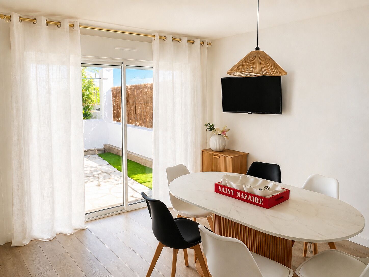 A modern dining area with a white table, black chairs, a red box labeled 'SAINT NAZARE,' and a TV on the wall.