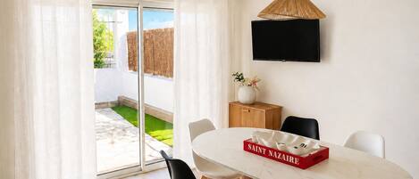 A modern dining area with a white table, black chairs, a red box labeled 'SAINT NAZARE,' and a TV on the wall.