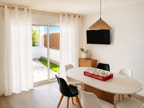 Uma área de refeições moderna com uma mesa branca, cadeiras pretas, uma caixa vermelha com a inscrição 'SAINT NAZARE' e uma TV na parede.