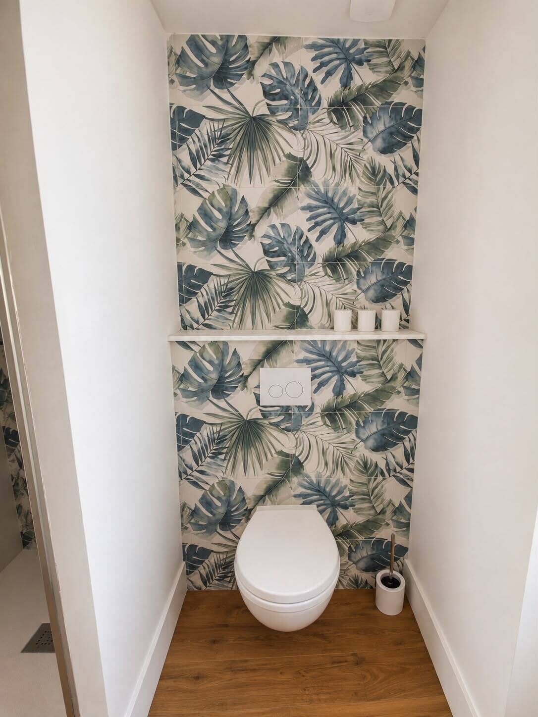 Et badeværelse med toilet, en hylde med to hvide beholdere, og en væg med et tropisk bladmotiv.