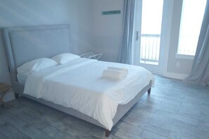 Una cama bien hecha con sábanas blancas y una toalla doblada, piso de madera y vistas al exterior a través de una puerta.