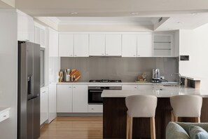 A modern kitchen with white cabinetry, a stainless steel refrigerator, a built-in oven, and a large island with bar stools. - Luxury 5BR canal retreat with private jetty, alfresco, water views & space for 10. (Dudley Park)