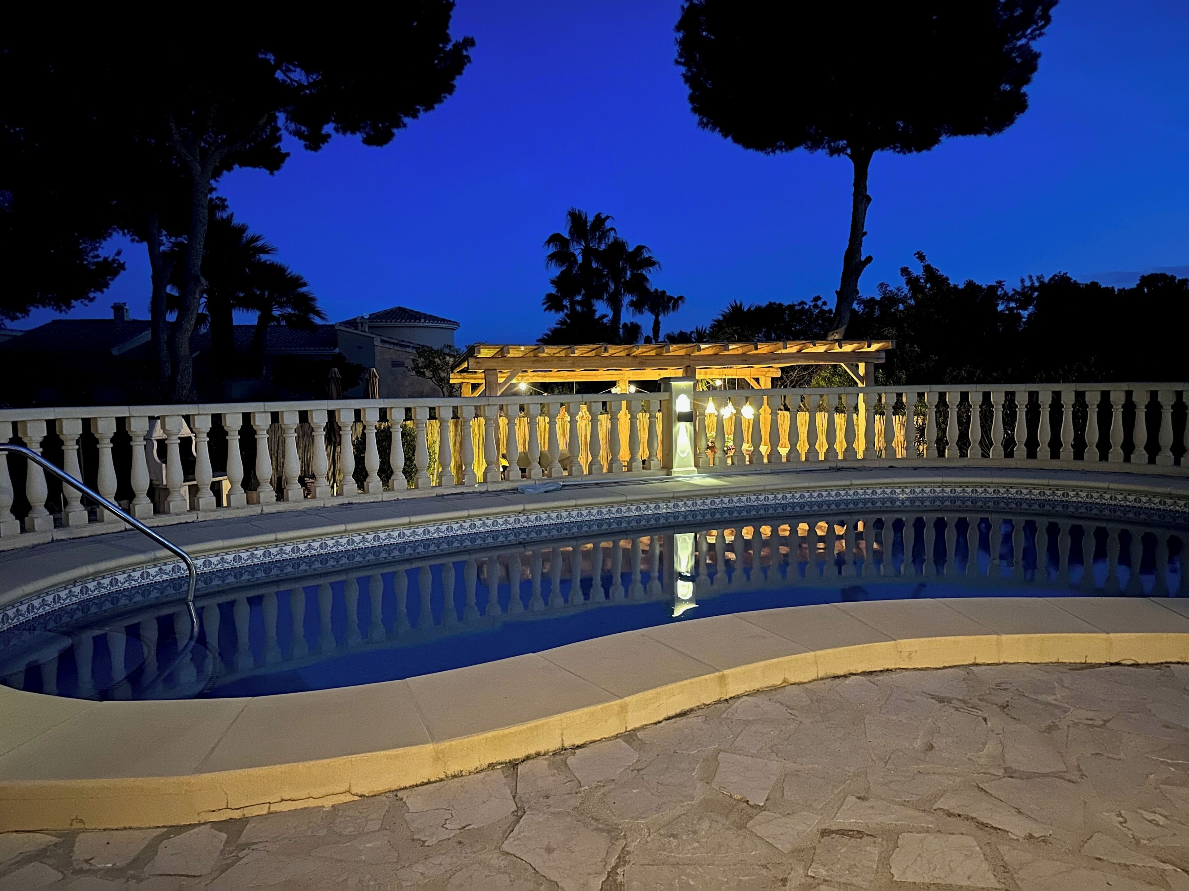 A swimming pool with a tiled edge, a white balustrade, and a building with illuminated windows in the background.