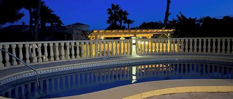 A swimming pool with a tiled edge, a white balustrade, and a building with illuminated windows in the background.