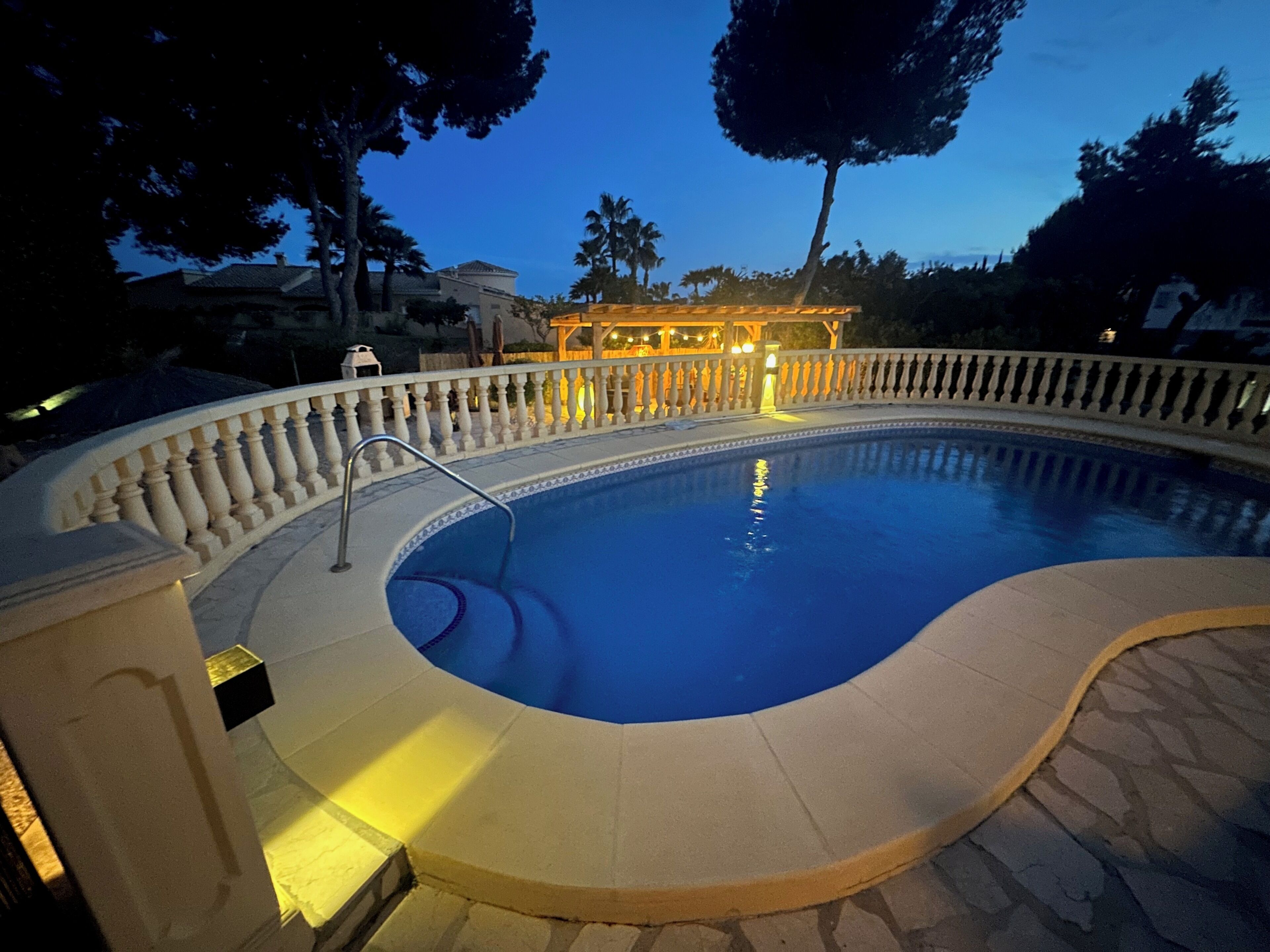 Une piscine à bord incurvé, un espace aménagé au bord de la piscine avec un garde-corps, et un bâtiment illuminé en arrière-plan.