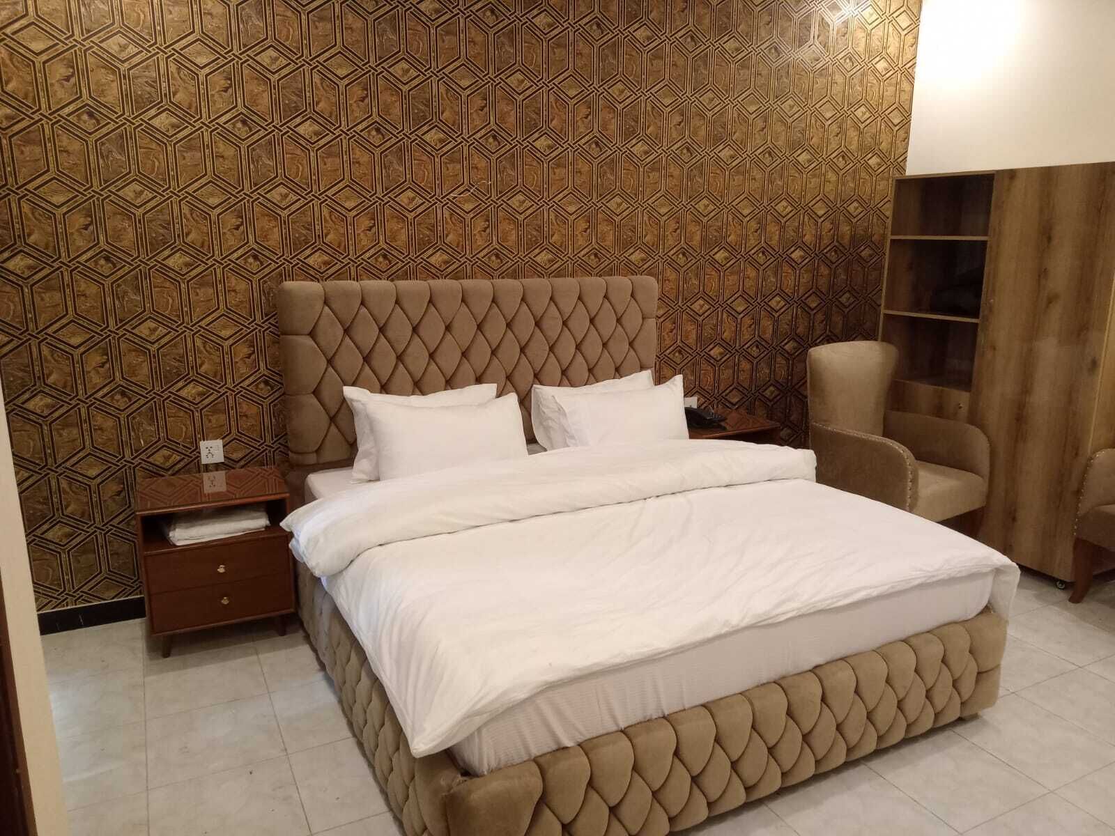 Quarto com cabeceira estofada, cama com roupa de cama branca, mesa de cabeceira, cadeira e prateleira de madeira.